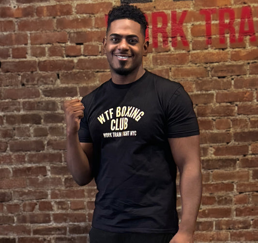 Man wearing a black 'WTF Boxing Club' t-shirt in front of a brick wall with 'WTF Train' sign